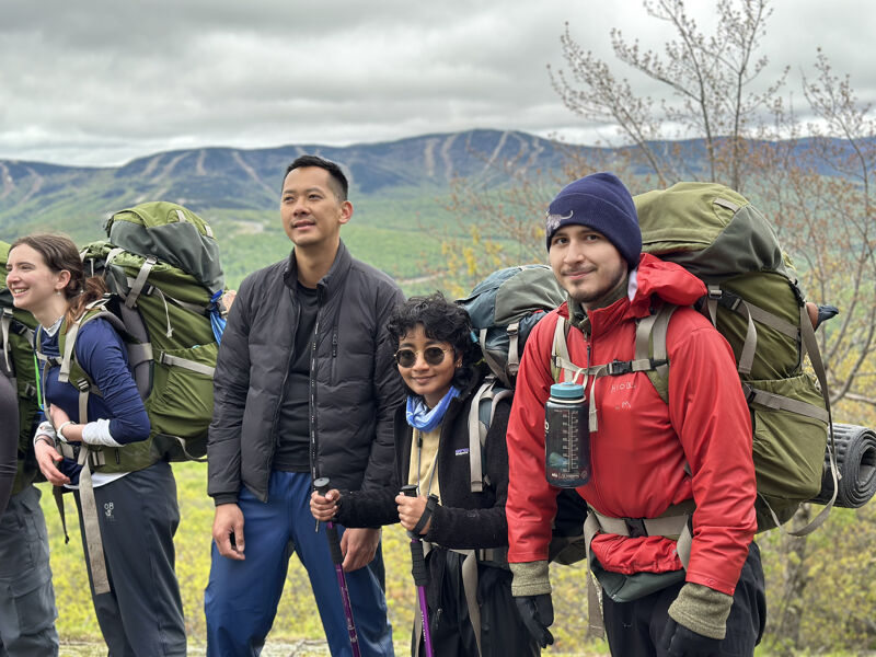 The image shows a group of five people on a hiking trip. They are standing on a rocky outcrop, with a scenic mountain range in the background. Each person is wearing hiking gear, including backpacks. The weather appears to be overcast, with a cloudy sky. The group seems to be taking a break to enjoy the view during their hike.